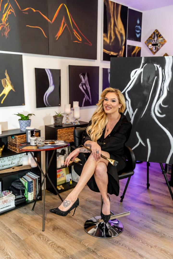 Ashley Karno sitting with a paintbrush in her studio, with paintings on the walls and a canvas beside her